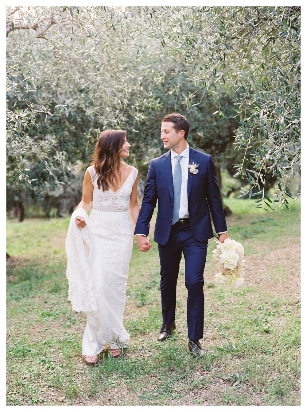  bride and groom wedding photos in olive grove in tuscany italy, villa le fontanelle wedding photos, tuscany wedding ideas, groom holding bouquet, bride bouquet roses, lace wedding dress fitted, florence wedding, florence wedding photos, tuscany wedding photography, bride and groom walking holding hands 